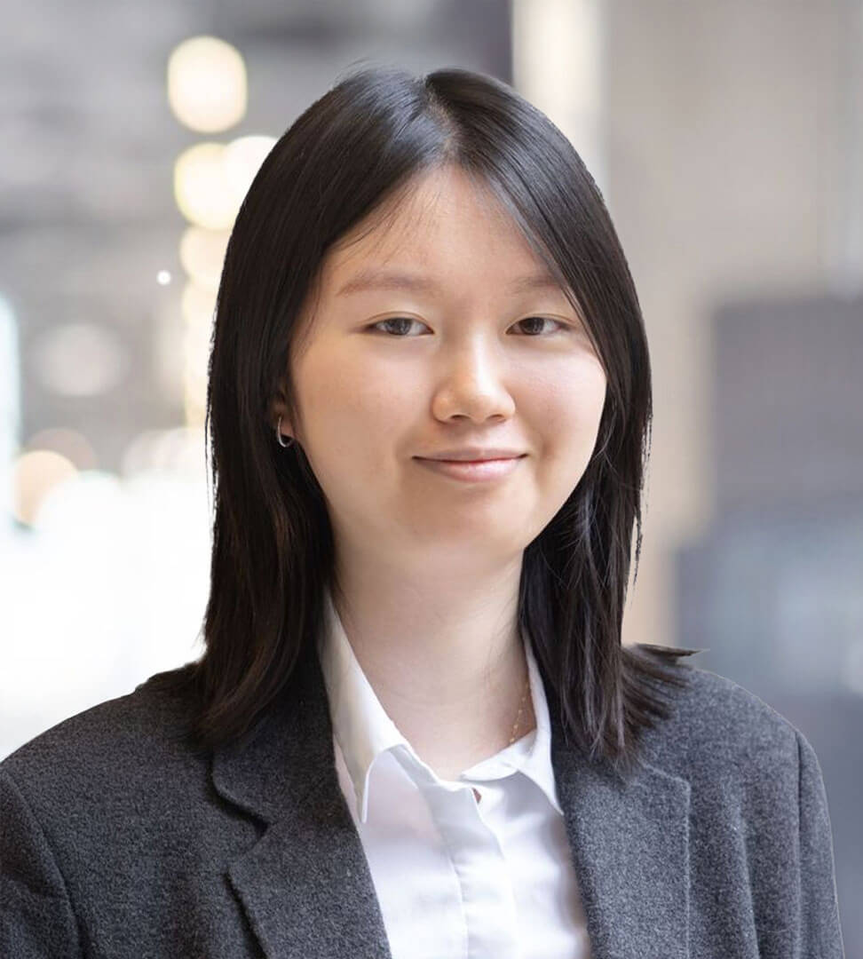 A young woman with shoulder-length black hair, wearing a white shirt and dark blazer, smiling softly in an indoor setting.