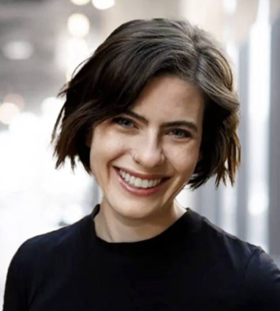A woman with short, wavy brown hair and a bright smile, wearing a black top, standing indoors with blurred lights in the