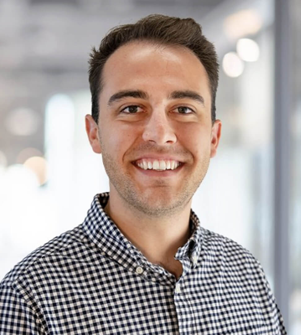 A smiling young man with short brown hair wearing a black-and-white checkered shirt, standing in a modern office environment.