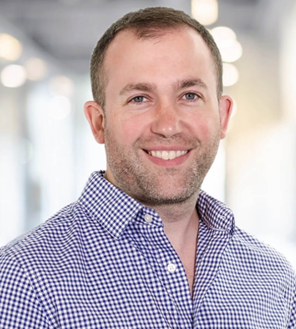 A smiling man with short brown hair, wearing a blue and white checkered shirt, in a bright, modern office environment.