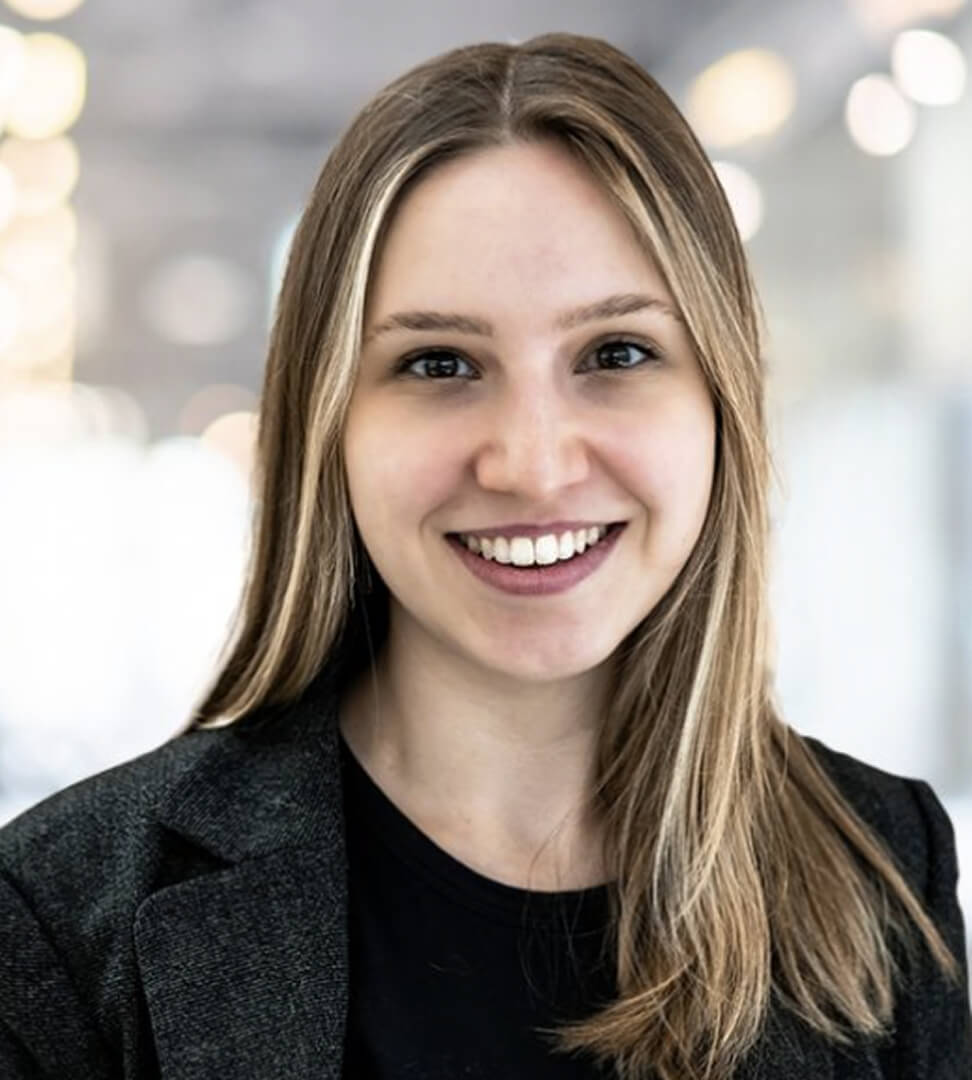 A smiling woman with long, light brown hair, wearing a dark blazer, in a professional setting with blurred lights in the