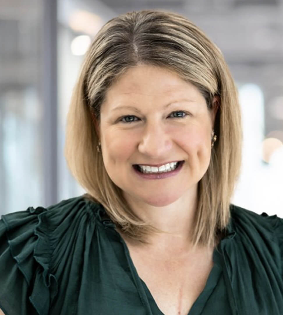 A smiling woman with shoulder-length blonde hair wearing a dark green top with ruffled shoulders.