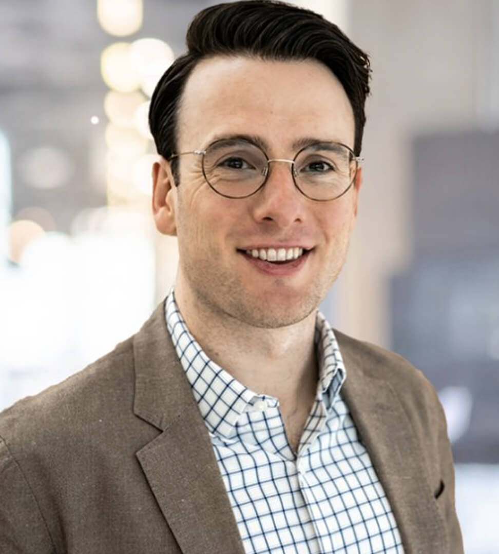 A smiling man with dark hair, glasses, wearing a checked shirt and brown blazer, in a professional indoor setting.