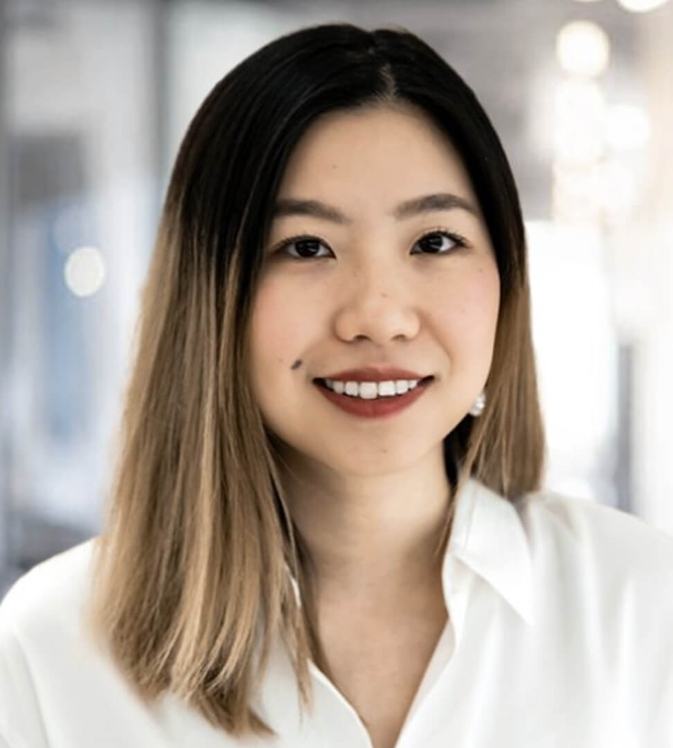 A woman with shoulder-length hair, wearing a white shirt, smiling in a bright, modern indoor setting.